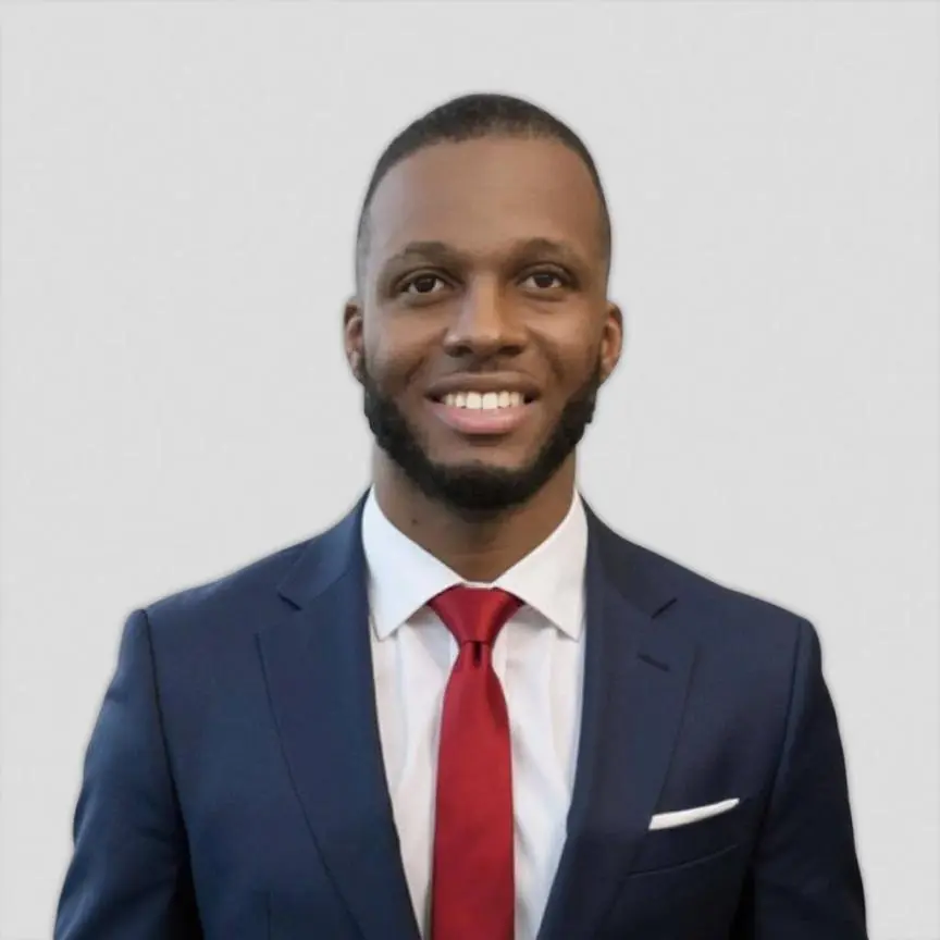 Smiling man in a navy suit, white shirt, red tie, and pocket square posing for a formal headshot against a light background.