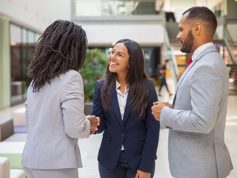 Three professionals greet with a handshake and smiles in a bright, modern office lobby.
