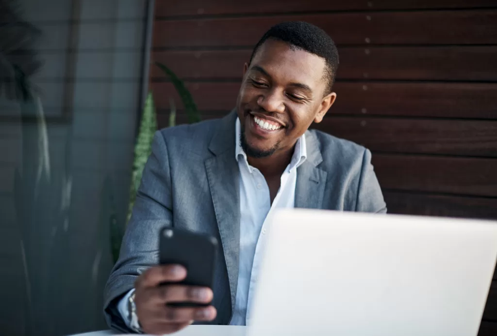 Smiling man in a gray blazer sits at a desk with a laptop and looks at his smartphone.
