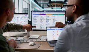 Two coworkers review a large monitor displaying a payroll dashboard in a bright office.