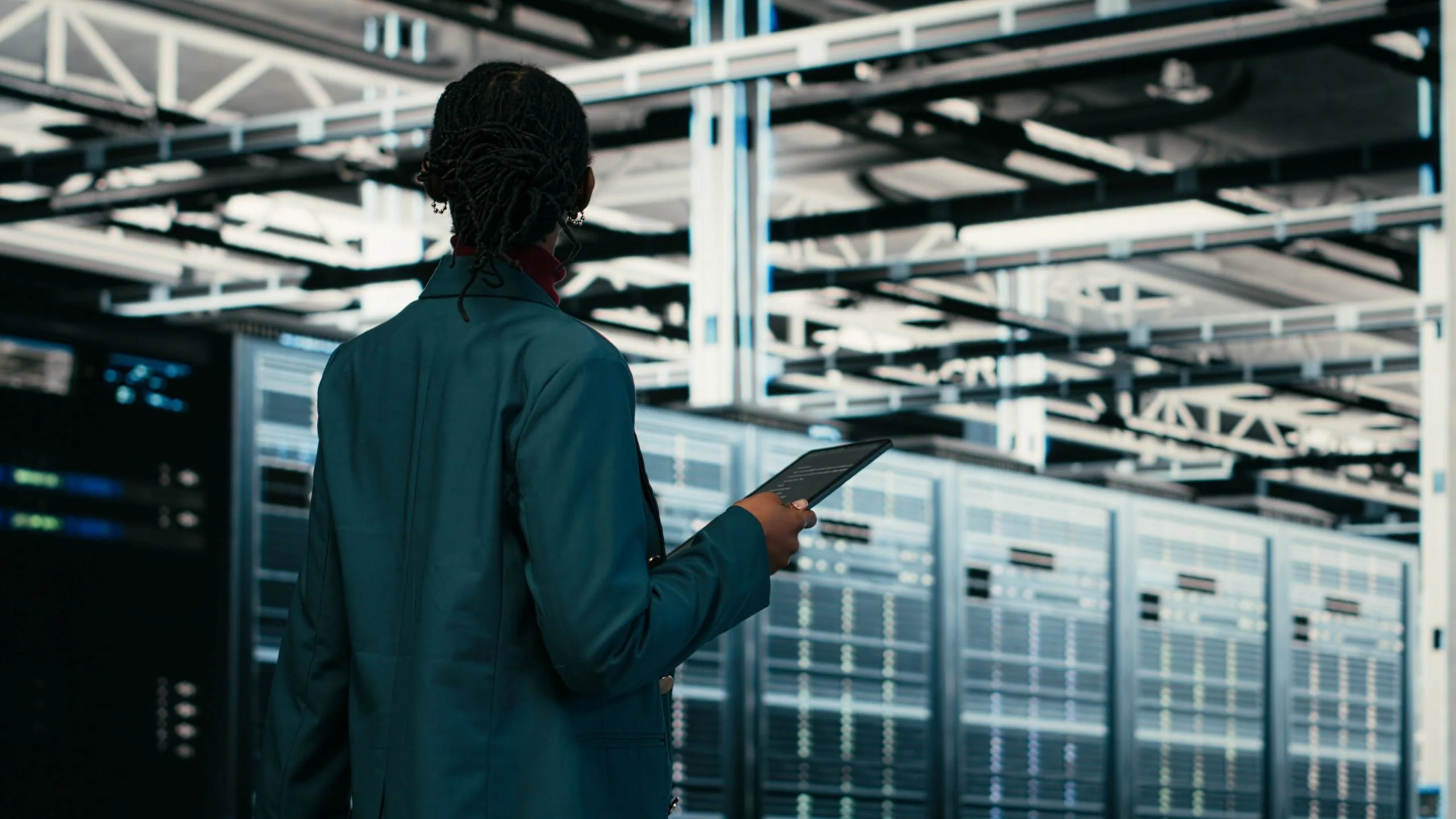 Person in a teal jacket stands with a tablet in a server room, facing racks of equipment and blinking lights on the side